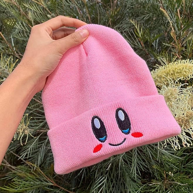 Pink Kirby Beanie with cartoon face design held by a hand against a green foliage background